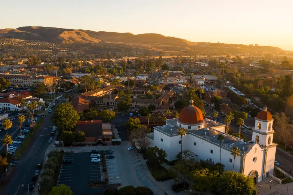 Aerial view of a small town at sunset, showing a white church with red domes and a bell tower in the foreground, palm trees and grid streets nearby.