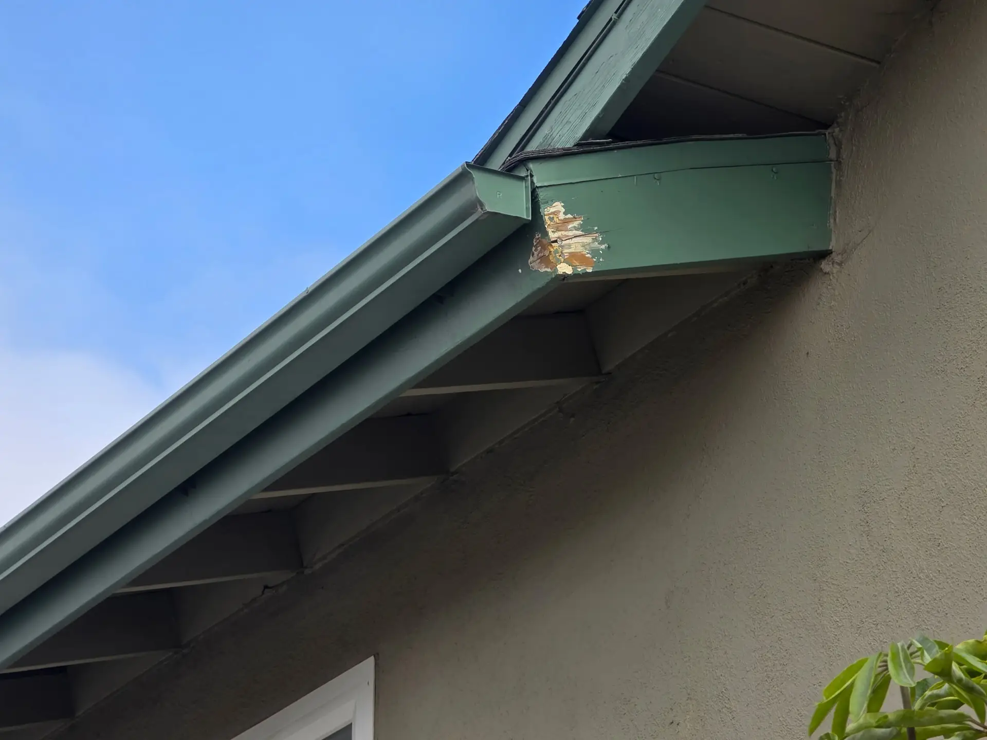 Green metal roof edge with peeling paint against a clear blue sky; part of an overhang and supporting beam visible.