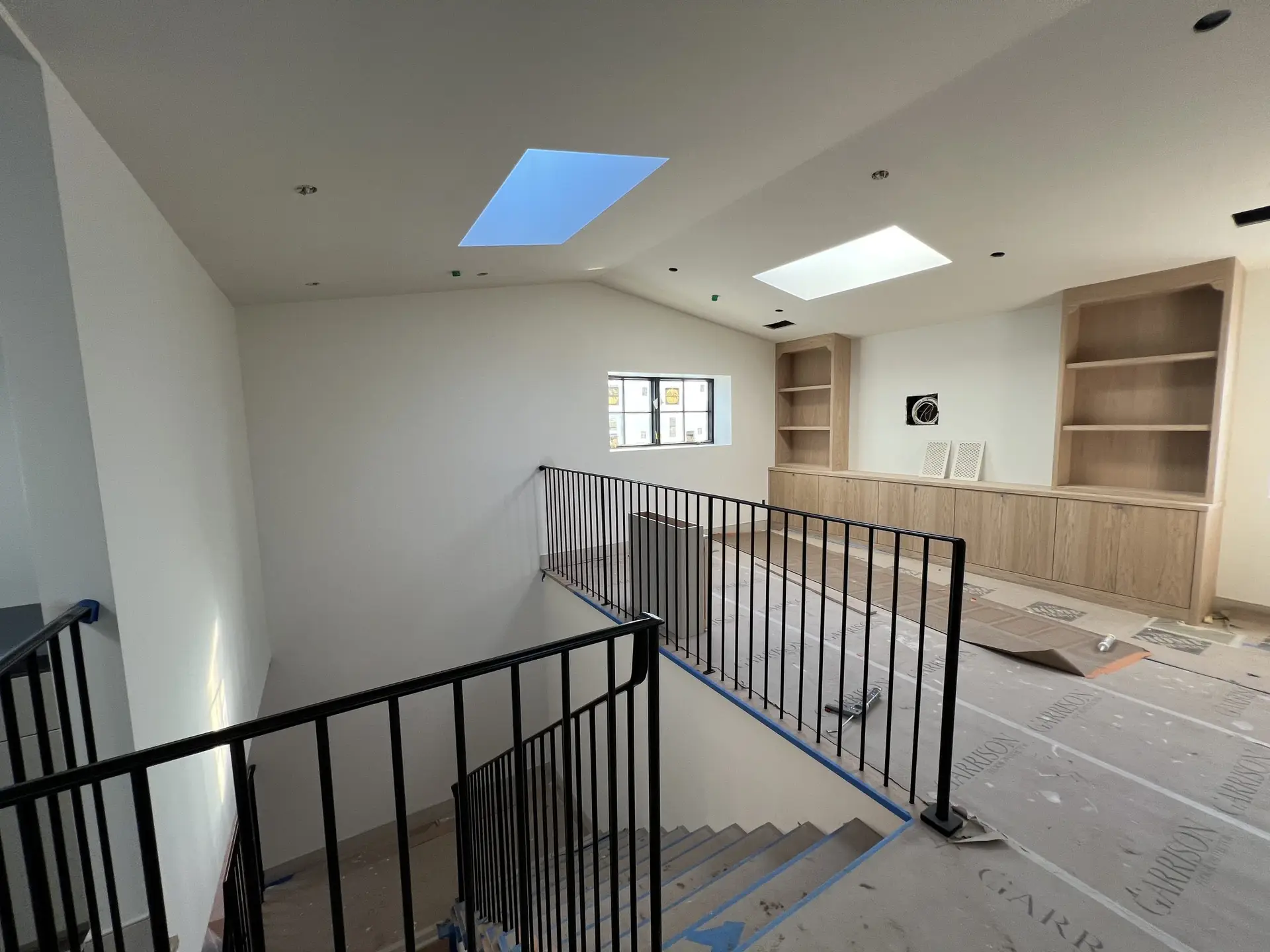 Interior stairwell under construction with black metal railings, light wood built-in shelves, and rectangular skylights in the ceiling.