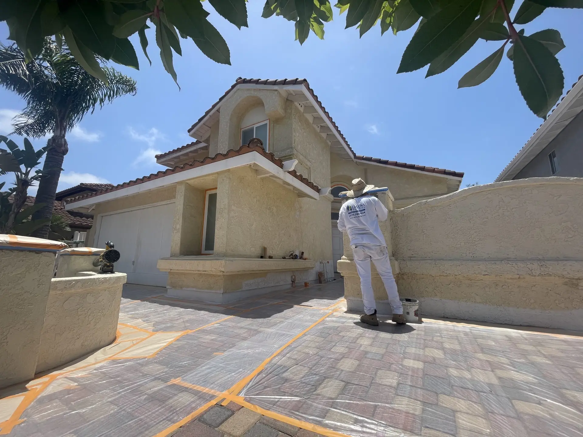 Construction worker in white protective clothing working on a beige stucco house with terracotta roof tiles under a clear blue sky, framed by palm trees. (informative)
