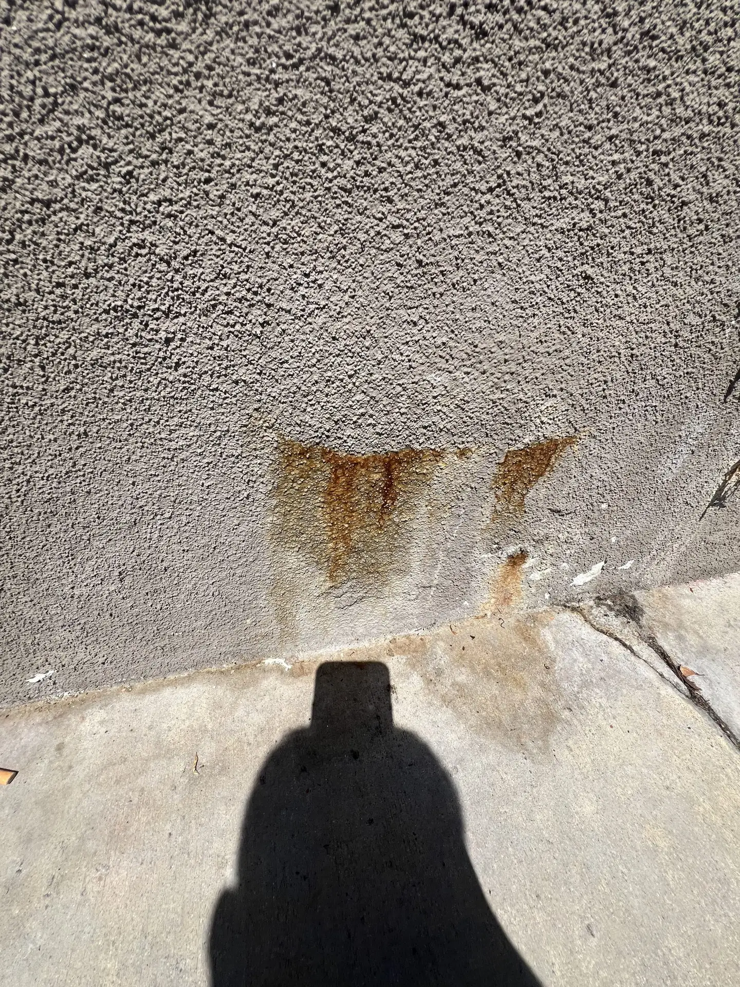 Shadow of a person taking a photo cast on a rough gray wall with rust stains above a concrete sidewalk.