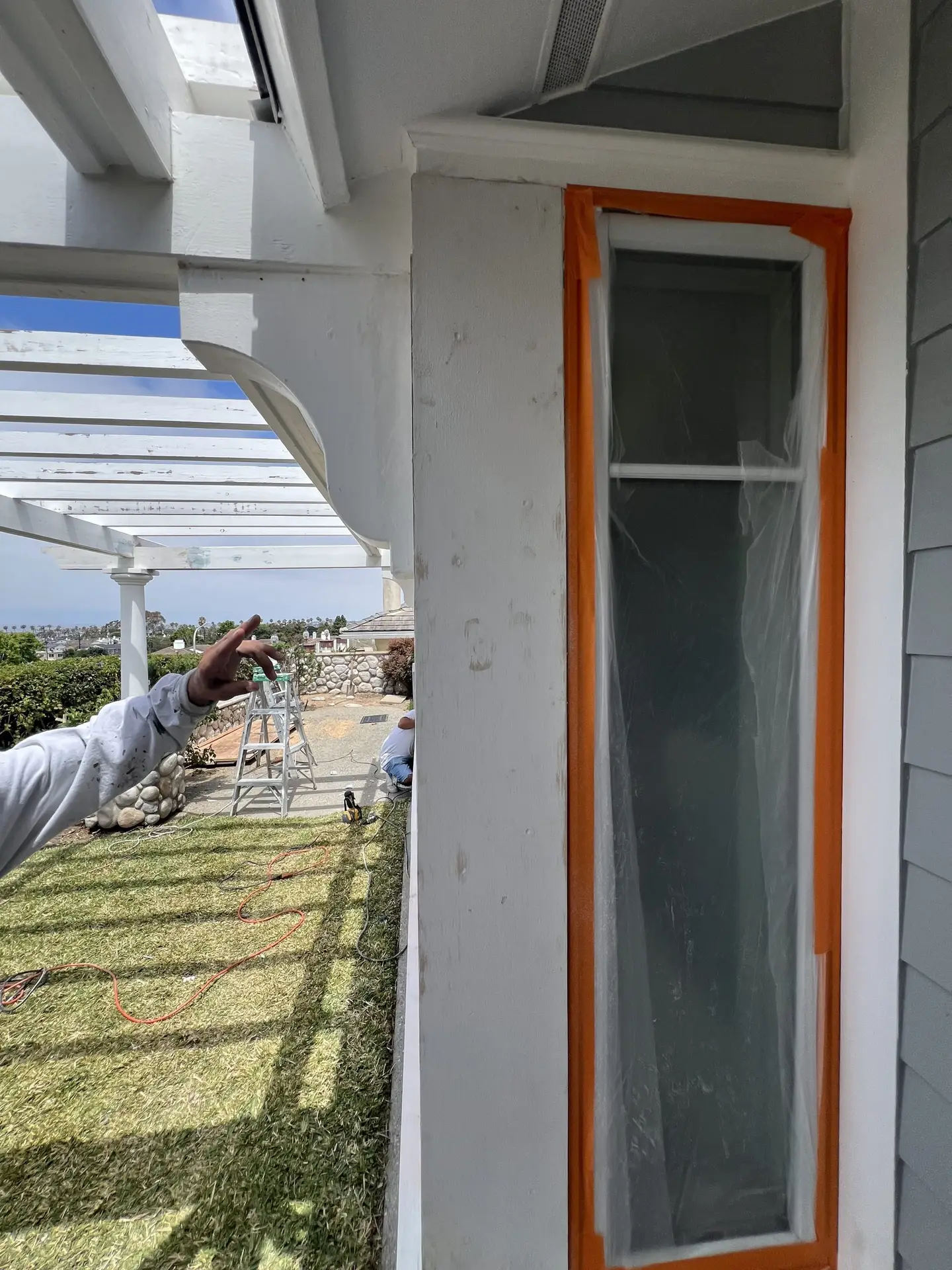 Exterior wall with a window frame outlined in orange tape; plastic on the glass, construction work underway near a white pergola.