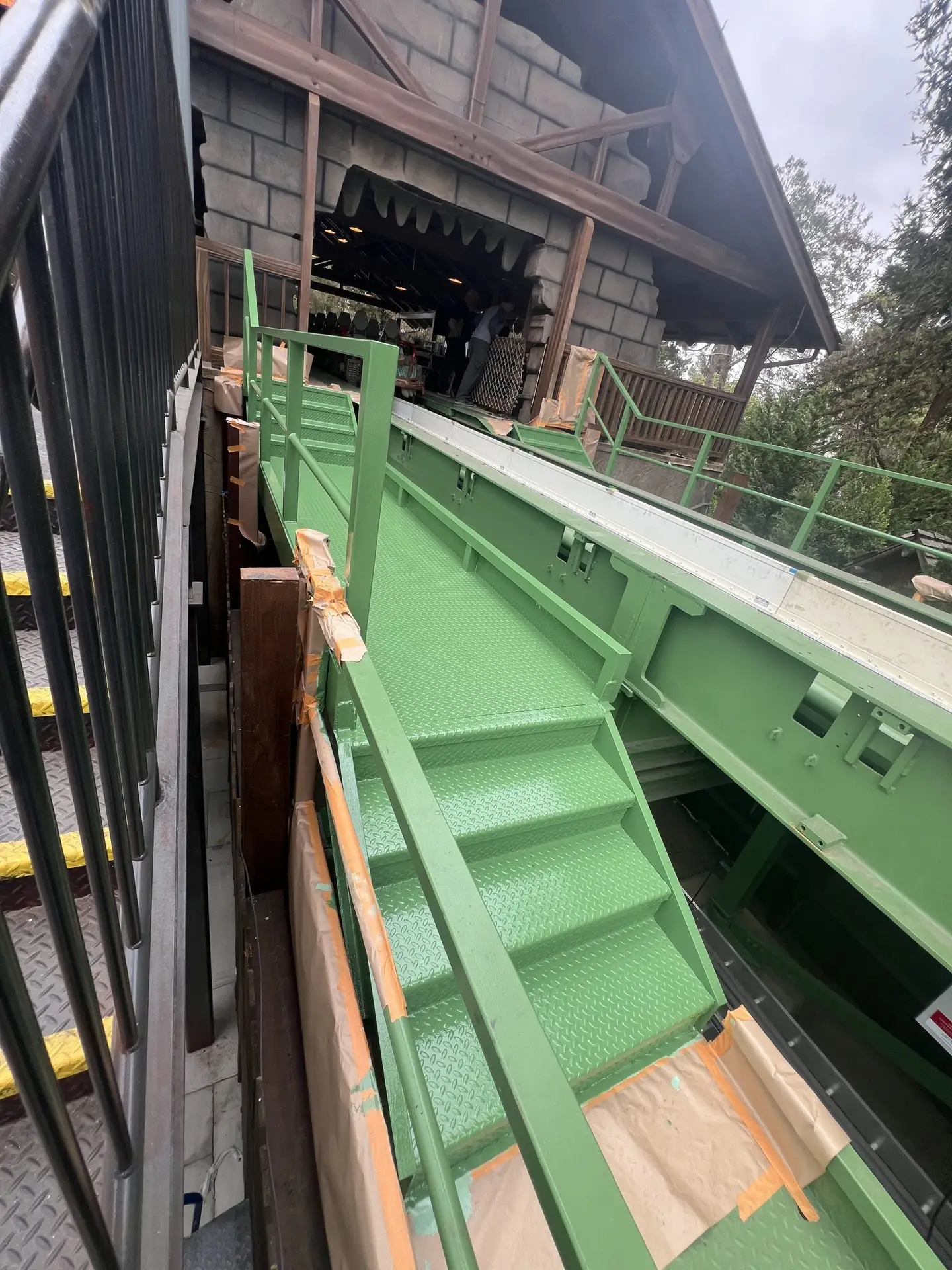 Green metal staircase and walkway along a building under construction, with protective cardboard edging and a worker near an open wall opening.