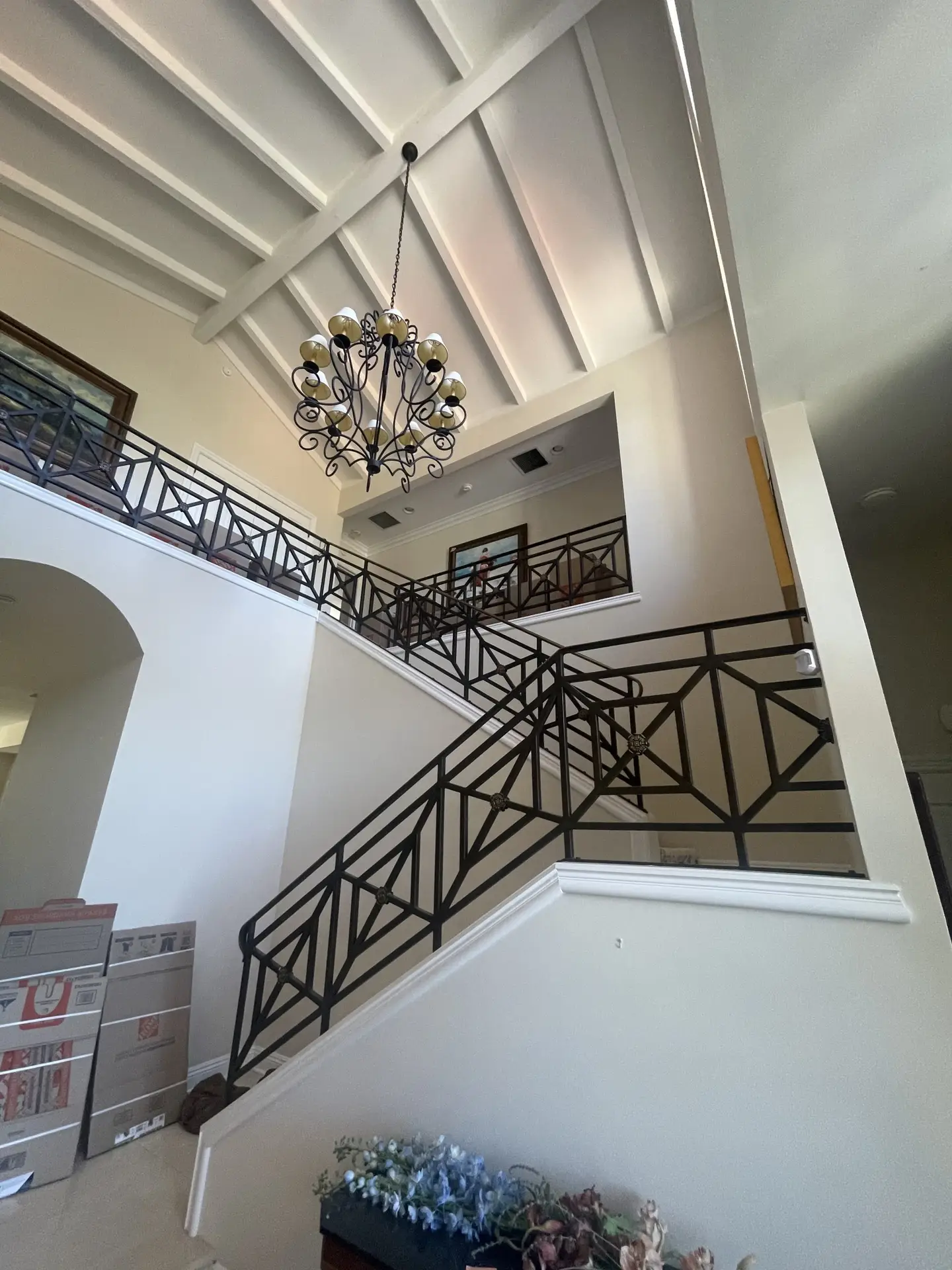 Two-story foyer with a black metal railing staircase, ornate chandelier, and exposed beam ceiling; boxed items along the left wall.