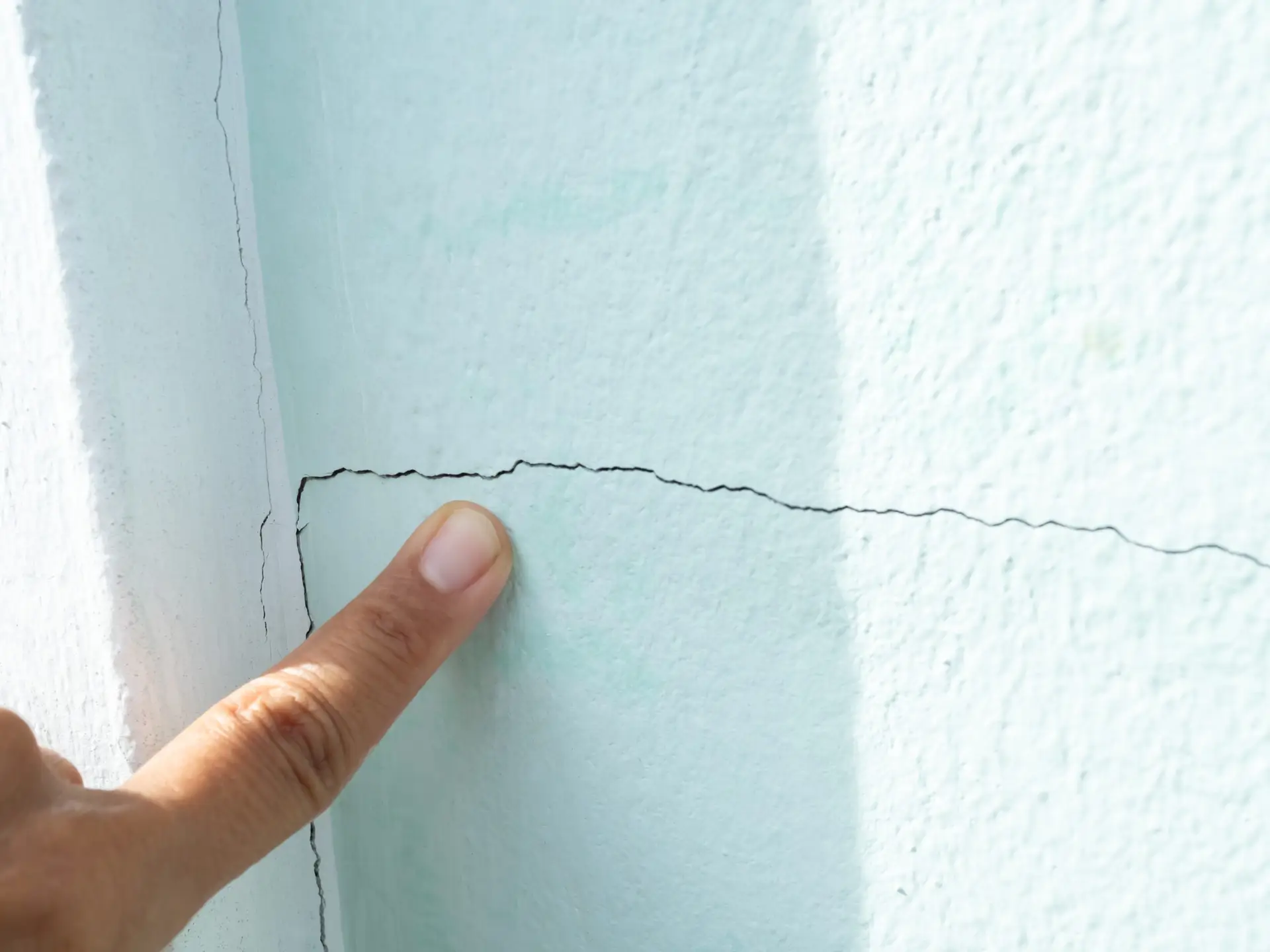Close-up of a finger pointing at a horizontal crack in a pale blue wall, highlighting wall damage.